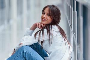 brune en chemise blanche est assise à l'intérieur dans un aéroport ou un couloir moderne pendant la journée photo