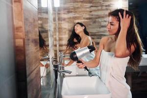 deux belles jeunes femmes debout dans la salle de bain près du miroir ensemble, se nettoyant le visage et séchant les cheveux photo