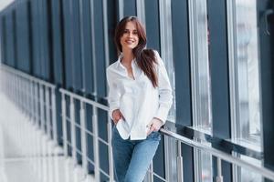 brune en chemise blanche s'appuyant sur des balustrades à l'intérieur d'un aéroport moderne ou d'un couloir pendant la journée photo