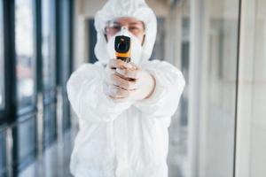 femme médecin scientifique en blouse de laboratoire, lunettes défensives et masque debout à l'intérieur avec thermomètre infrarouge photo