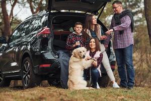 une famille heureuse s'amuse avec son chien près d'une voiture moderne à l'extérieur dans la forêt photo