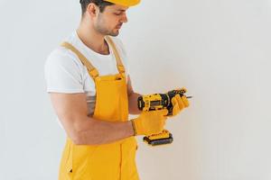 homme à tout faire en uniforme jaune debout contre un mur blanc avec un tournevis automatique. conception de rénovation de maison photo