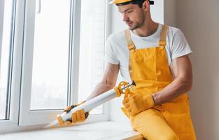 homme à tout faire en uniforme jaune travaille avec de la colle pour fenêtre à l'intérieur. conception de rénovation de maison photo