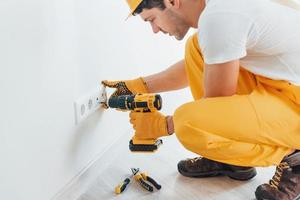 homme à tout faire en uniforme jaune travaille avec l'électricité et installe une nouvelle prise à l'aide d'un tournevis automatique. conception de rénovation de maison photo