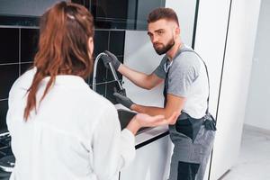 un jeune plombier professionnel en uniforme gris montre les résultats du travail d'une femme au foyer dans la cuisine photo