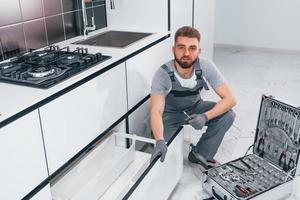 jeune plombier professionnel en uniforme gris utilisant du matériel dans la cuisine et se préparant au travail photo