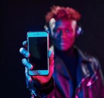 avec téléphone et écouteurs. éclairage néon futuriste. jeune homme afro-américain dans le studio photo