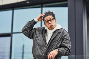 beau jeune homme aux cheveux noirs bouclés et à lunettes est dans la rue contre le bâtiment photo