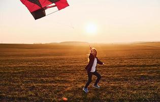 bonne petite fille qui court avec un cerf-volant dans les mains sur le beau terrain au moment du soleil photo