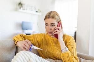 jeune femme regarde le thermomètre. elle a de la fièvre, elle appelle son médecin. belle femme avec des symptômes appelée numéro d'urgence covid-19 pour signaler ses conditions médicales photo