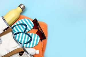 accessoires de plage - une bouteille avec une boisson non alcoolisée, de la crème solaire, des lunettes de soleil, des tongs, une serviette dans un sac de plage sur fond bleu avec espace de copie photo