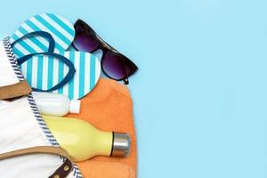 accessoires de plage - une bouteille avec une boisson non alcoolisée, de la crème solaire, des lunettes de soleil, des tongs, une serviette dans un sac de plage sur fond bleu avec espace de copie photo