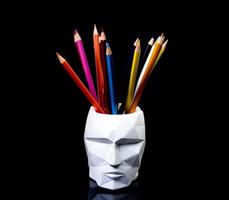 des crayons de couleur pour le dessin, situés dans un support comme un vase, se concentrent devant. crayons multicolores et feutres dans un vase en plâtre en forme de visage sur fond noir. photo