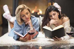 deux amies lisant un livre et buvant du vin rouge au coin du feu. concept de style de vie. photo