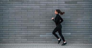 jeune fille sportive en tenue de sport noire courant à l'extérieur près d'un mur gris photo