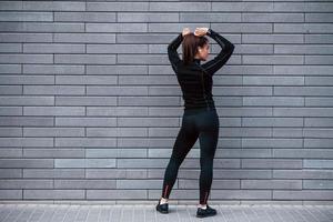 vue arrière d'une jeune fille sportive en tenue de sport noire qui se tient à l'extérieur près d'un mur gris photo