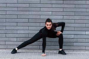 jeune fille sportive en tenue de sport noire à l'extérieur faisant des étirements près d'un mur gris photo