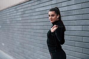 jeune fille sportive en tenue de sport noire debout avec une corde à sauter dans les mains à l'extérieur près d'un mur gris photo