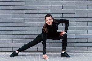 jeune fille sportive en tenue de sport noire à l'extérieur faisant des étirements près d'un mur gris photo