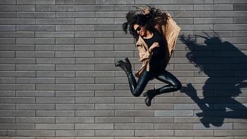 belle brune aux cheveux bouclés et en vêtements noirs sautant et s'amusant à l'extérieur près du mur photo