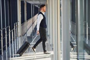 un jeune passager masculin vêtu d'élégants vêtements de cérémonie se trouve dans le hall de l'aéroport en train de marcher avec des bagages photo