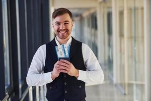 jeune passager masculin dans des vêtements formels élégants souriant et tenant des billets dans les mains dans le hall de l'aéroport photo