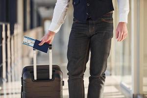 vue rapprochée d'un passager masculin vêtu d'élégants vêtements formels qui se trouve dans le hall de l'aéroport avec des bagages et des billets photo