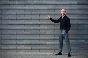 homme élégant à la mode debout à l'extérieur contre un mur gris et montre à gauche par le doigt photo