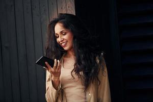 femme aux cheveux bouclés noirs debout contre l'extérieur du bâtiment en bois noir et utilisant le téléphone photo