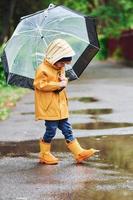 enfant en cape imperméable jaune, bottes et parapluie jouant à l'extérieur après la pluie photo