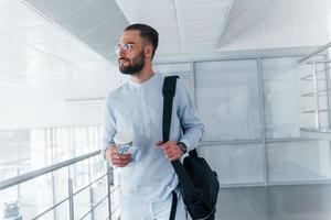 tenant une tasse avec boisson et sac noir. jeune bel homme en vêtements formels à l'intérieur du bureau pendant la journée photo