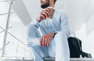 tenant des billets d'avion à la main. assis dans les escaliers et attendant. jeune passager en tenue de soirée à l'intérieur du bureau pendant la journée photo