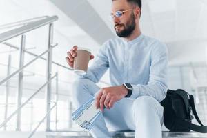 tenant des billets d'avion à la main. assis dans les escaliers et attendant. jeune passager en tenue de soirée à l'intérieur du bureau pendant la journée photo