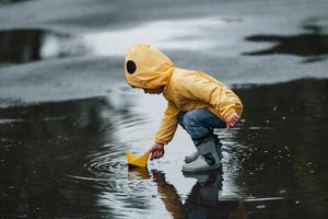 enfant en cape imperméable jaune et bottes jouant avec un jouet de bateau fait main en papier à l'extérieur après la pluie photo