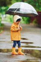 enfant en cape imperméable jaune, bottes et parapluie jouant à l'extérieur après la pluie photo