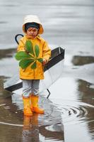 enfant en cape imperméable jaune, bottes et parapluie jouant à l'extérieur après la pluie photo