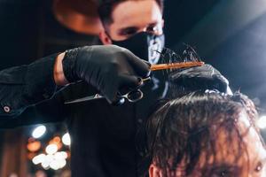 jeune homme barbu assis et se faisant couper les cheveux dans un salon de coiffure par un gars en masque de protection noir photo