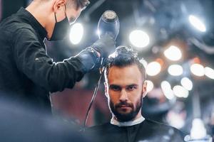 vue de face d'un jeune homme barbu assis et se faisant couper les cheveux dans un salon de coiffure par un gars en masque de protection noir photo