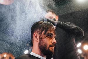 jeune homme barbu assis et se faisant couper les cheveux dans un salon de coiffure par un gars en masque de protection noir photo