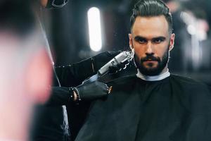 jeune homme avec une coiffure élégante assis et se rasant la barbe dans un salon de coiffure photo