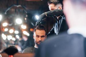 vue de face d'un jeune homme barbu assis et se faisant couper les cheveux dans un salon de coiffure par un gars en masque de protection noir photo