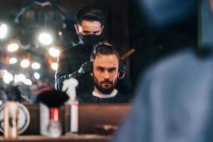 vue de face d'un jeune homme barbu assis et se faisant couper les cheveux dans un salon de coiffure par un gars en masque de protection noir photo