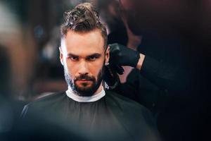 vue de face d'un jeune homme barbu assis et se faisant couper les cheveux dans un salon de coiffure par un gars en masque de protection noir photo