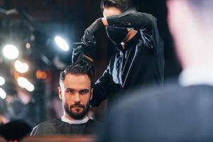 vue de face d'un jeune homme barbu assis et se faisant couper les cheveux dans un salon de coiffure par un gars en masque de protection noir photo