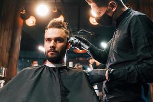 vue de face d'un jeune homme barbu assis et se faisant couper les cheveux dans un salon de coiffure par un gars en masque de protection noir photo