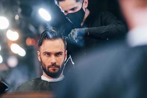 vue de face d'un jeune homme barbu assis et se faisant couper les cheveux dans un salon de coiffure par un gars en masque de protection noir photo