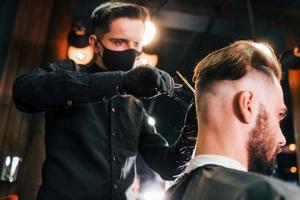 jeune homme barbu assis et se faisant couper les cheveux dans un salon de coiffure par un gars en masque de protection noir photo