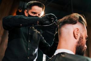 jeune homme barbu assis et se faisant couper les cheveux dans un salon de coiffure par un gars en masque de protection noir photo