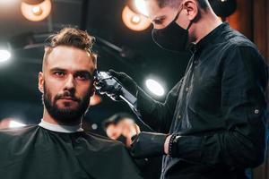 vue de face d'un jeune homme barbu assis et se faisant couper les cheveux dans un salon de coiffure par un gars en masque de protection noir photo