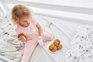 prendre un petit déjeuner en mangeant des biscuits. jolie petite fille en vêtements décontractés est à l'intérieur à la maison pendant la journée photo